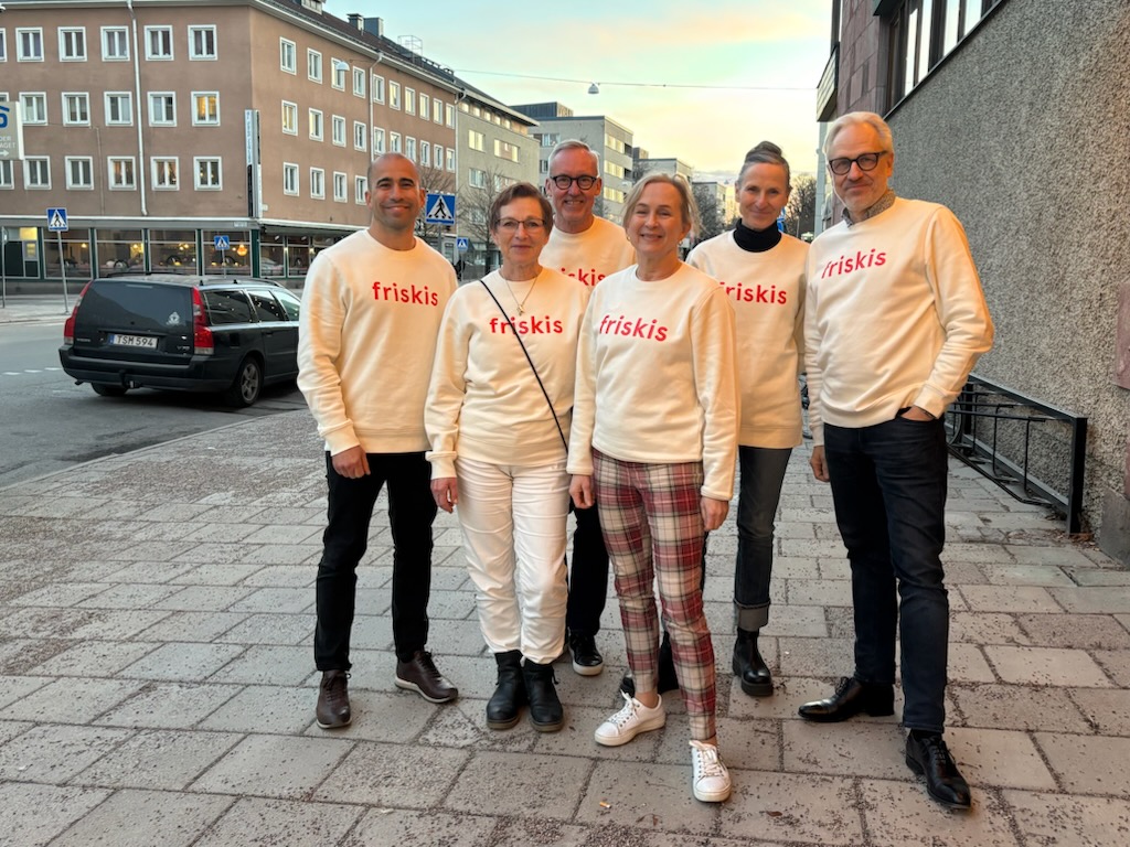 6 st personer står utomhus på en trottoar, de har vita tröjor med texten Friskis på bröstet. 